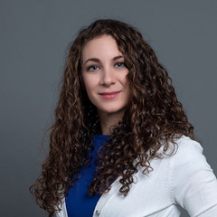 Rachael is a white woman with long curly dark brown hair and blue eyes, she wears a blue dress and white cardigan